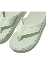 Fitflop Surff Webbing Toe Post Sandals. A pair of sage green sandals with cushioned sole, wedge heel, and wide patterned strap.