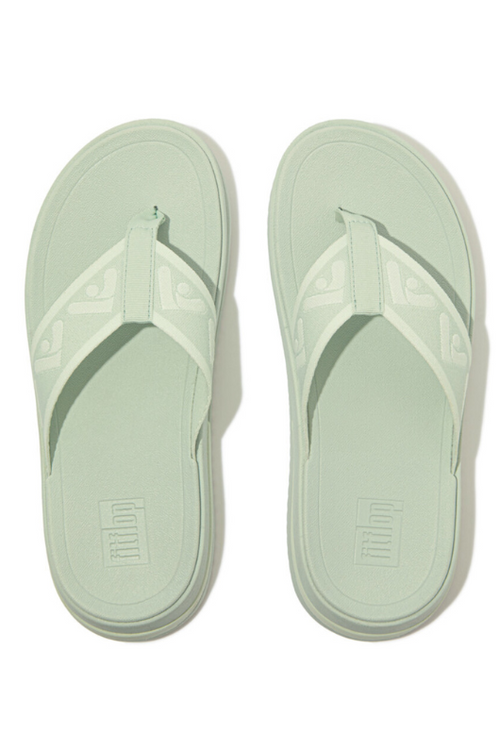 Fitflop Surff Webbing Toe Post Sandals. A pair of sage green sandals with cushioned sole, wedge heel, and wide patterned strap.