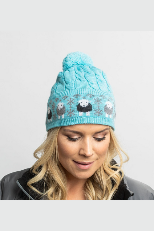 An image of a model wearing the Herdy Company Cable Knit Bobble Hat in Blue with a pom-pom and sheep design.