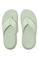 Fitflop Surff Webbing Toe Post Sandals. A pair of sage green sandals with cushioned sole, wedge heel, and wide patterned strap.