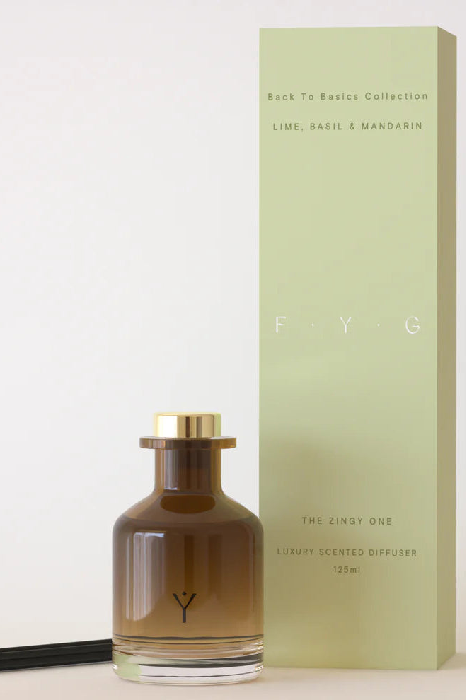 A luxury scented diffuser bottle with a gold cap and black reed stick, standing next to its pale green box on a light beige background. The bottle is amber-brown glass with a gold monogram logo.