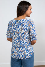 Lily & Me Meadow Top Confetti. A regular fit, feminine top with elbow-length sleeves, a feminine round neckline, curved hem, and a summery print with a blue and tan background, and white flower detail.