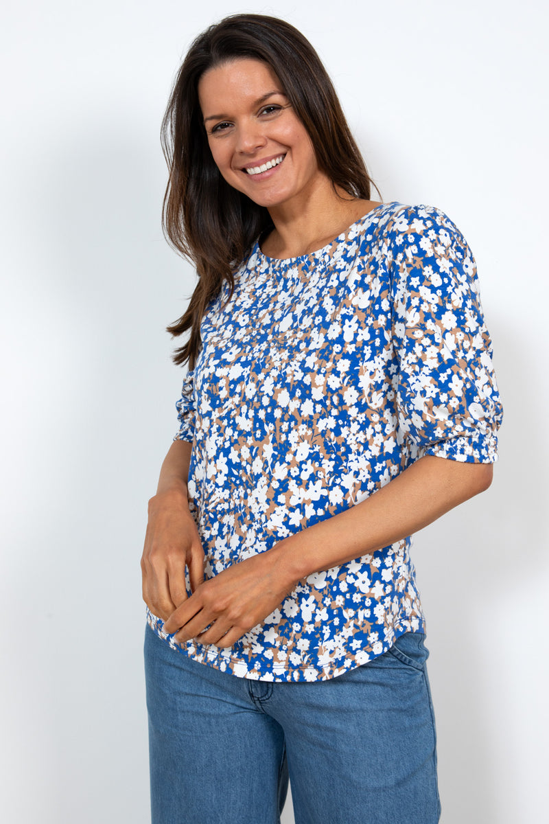 Lily & Me Meadow Top Confetti. A regular fit, feminine top with elbow-length sleeves, a feminine round neckline, curved hem, and a summery print with a blue and tan background, and white flower detail.