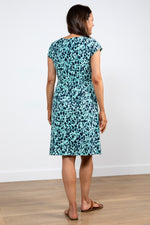 Lily & Me Harbourside Dress Confetti. A knee-length dress with short cap sleeves, a feminine round neckline, inverted pleats at the waist, and a turquoise design with black flower detail.
