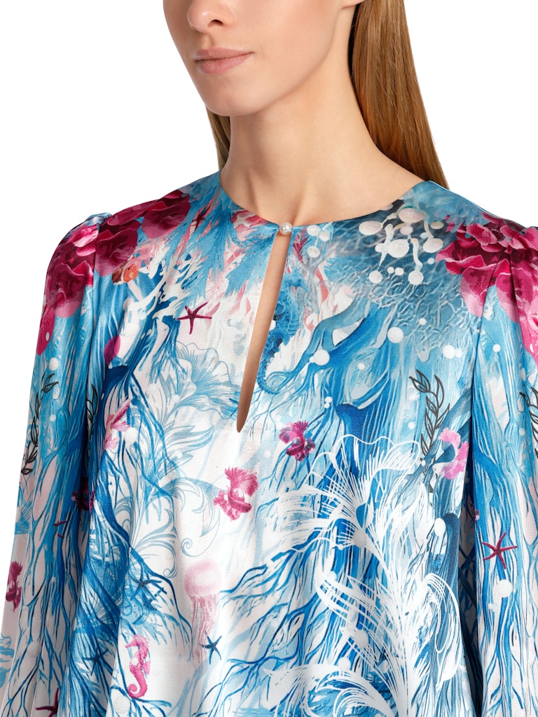 Marc Cain Long Sleeve Pattern Blouse. A relaxed fit blouse with long sleeves, featuring a ruffle detail. This top has a round neck with slit detail and back zip. The design has an underwater themed print in a mix of blue and red.