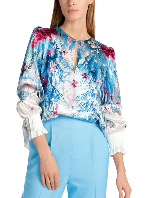 Marc Cain Long Sleeve Pattern Blouse. A relaxed fit blouse with long sleeves, featuring a ruffle detail. This top has a round neck with slit detail and back zip. The design has an underwater themed print in a mix of blue and red.